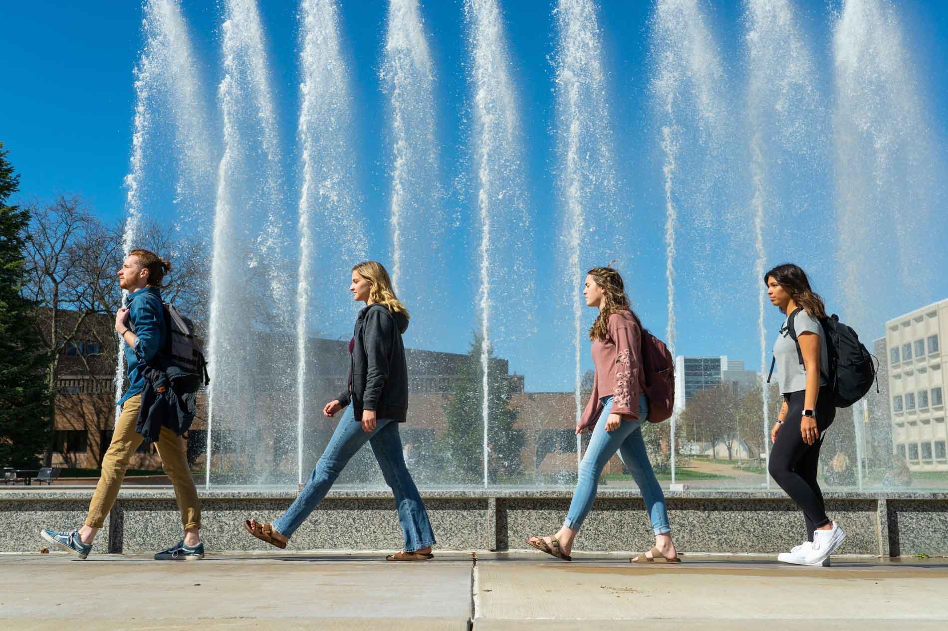 students walk in front of a fountain on campus